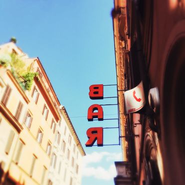 A color photo taken in Rome, Italy, of a red sign against a blue sky. The sign says BAR but has been taken from behind the sign so the letters are flipped.
